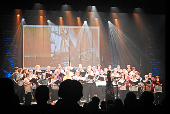 Un grand groupe de personnes chante sur une scène devant un grand écran.