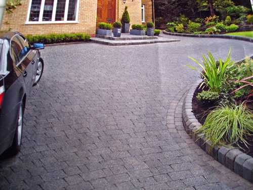 Dark gray brick driveway leading to a house with a car parked on the left and landscaping on the right.