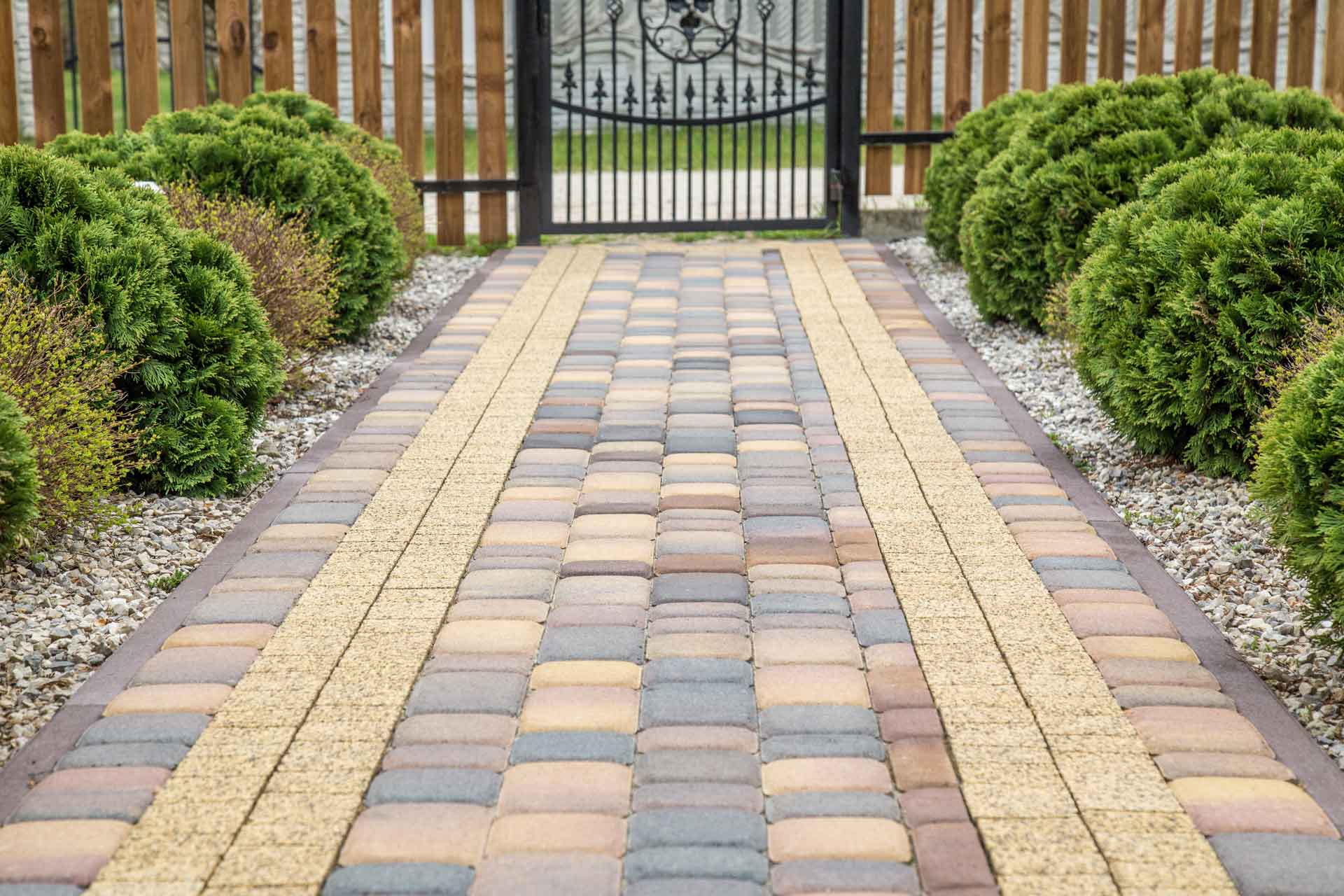 Brick pathway leading to a gate, bordered by green bushes and a wooden fence.