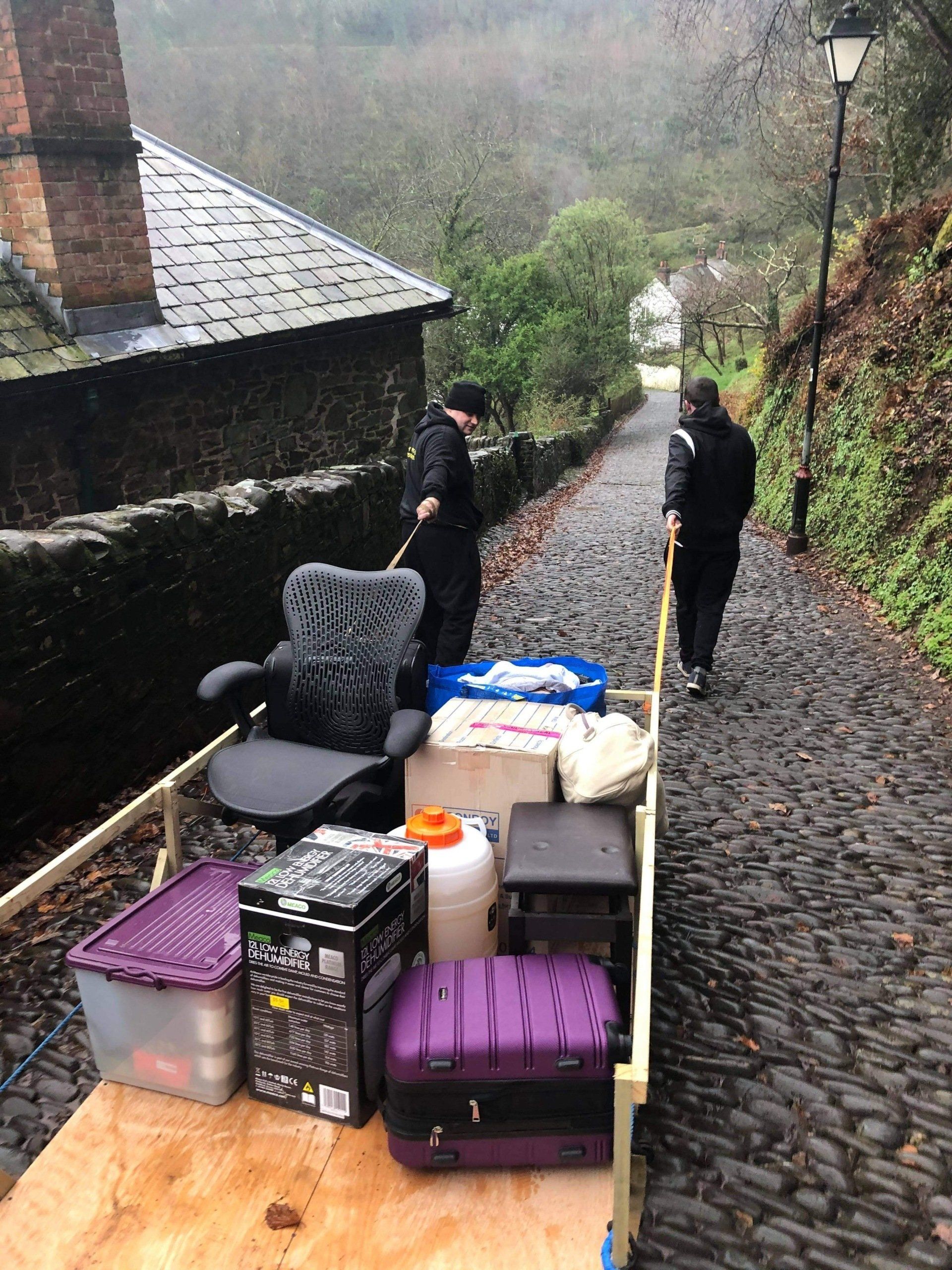 Two people on a narrow, wet path beside a stone wall, with stacked luggage and bags in the foreground.