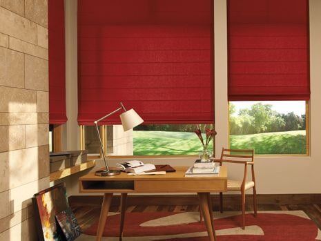 A room with a desk , chair , lamp and red blinds.
