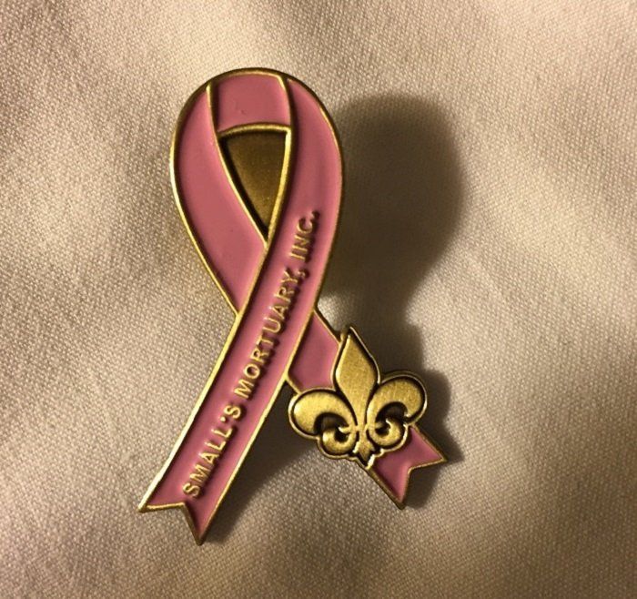 A pink awareness ribbon pin with a gold fleur-de-lis, inscribed with 