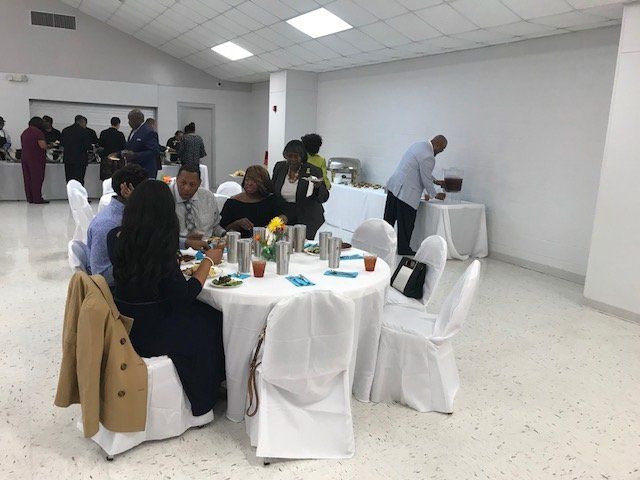 People gather at round tables in a bright, white-walled event space for a catered meal or social gathering.