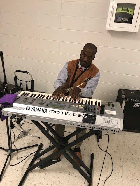 A person wearing a brown vest over a light shirt playing a Yamaha Motif ES6 keyboard on a stand indoors.