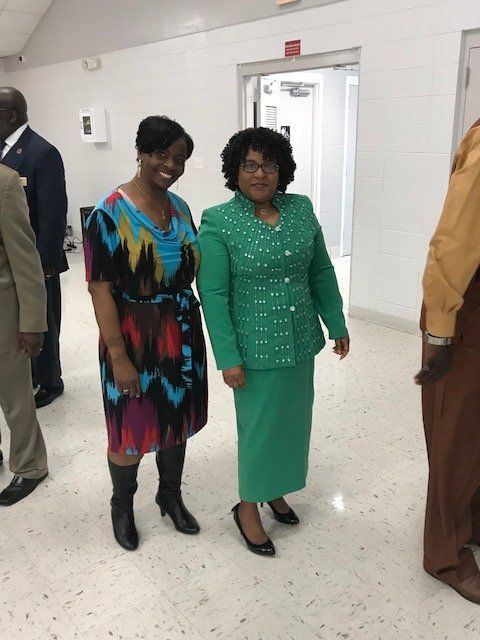 Two women smile while standing indoors. One wears a patterned dress and boots; the other wears a green suit and heels.