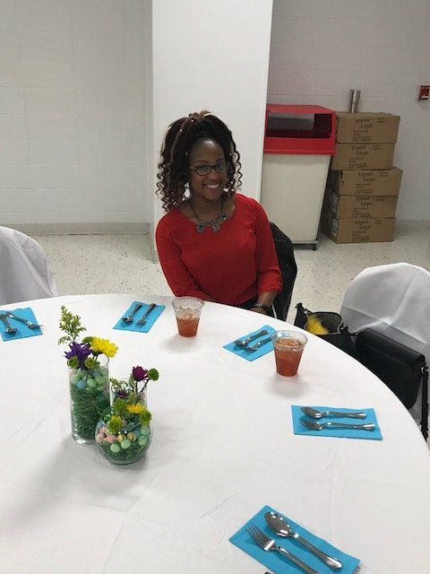 A person wearing a red shirt smiles while sitting at a round table set with colorful flowers and blue cutlery settings.