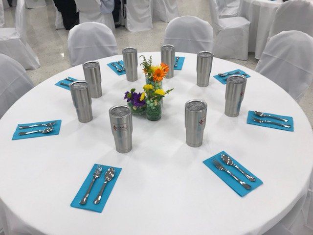 A round table with white tablecloth, set with stainless steel tumblers and blue napkins with silverware for each guest.
