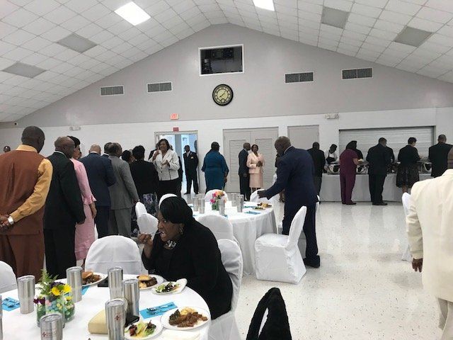 People gathered for a meal in a large, brightly lit hall with round tables and white tablecloths.
