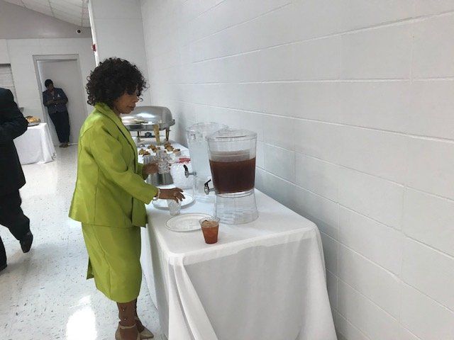 A person in a lime green suit pours a dark beverage from a dispenser at a table covered with a white cloth.