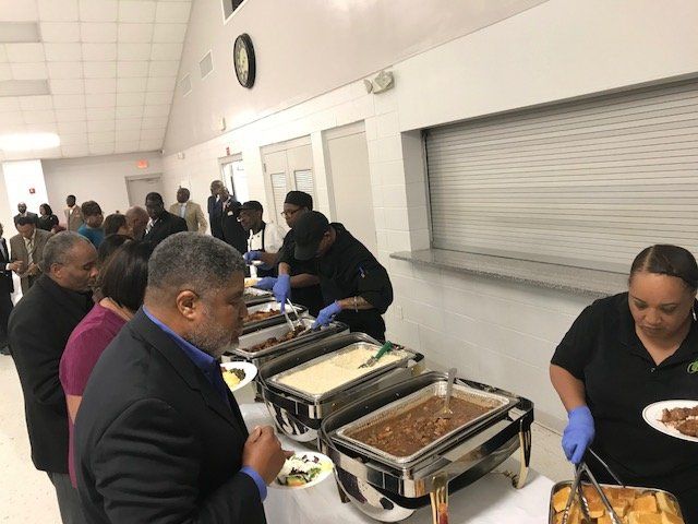 People wait in a buffet line in a dining hall, where staff serve food from metal chafing dishes on a long white table.