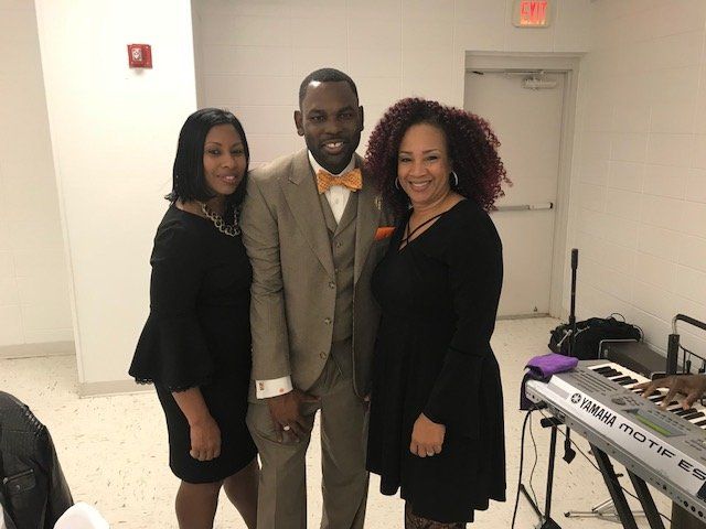 Three people pose for a photo in a room with a keyboard; a man in a tan suit stands between two women in black dresses.