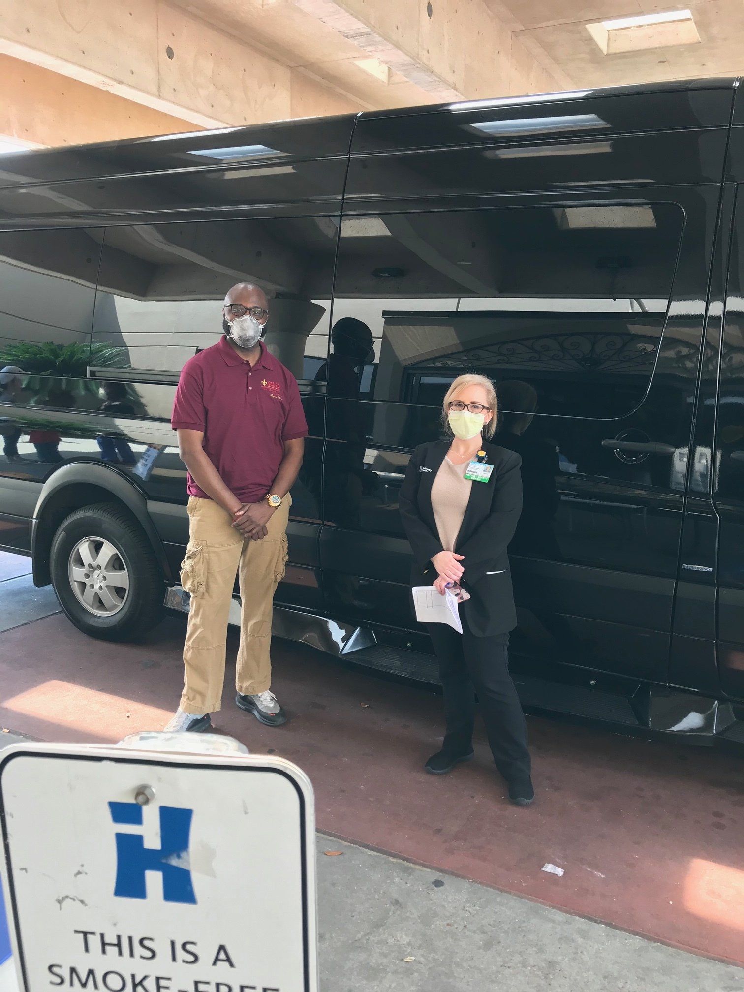 Two people wearing masks stand in front of a black vehicle under a parking structure next to a smoke-free sign.