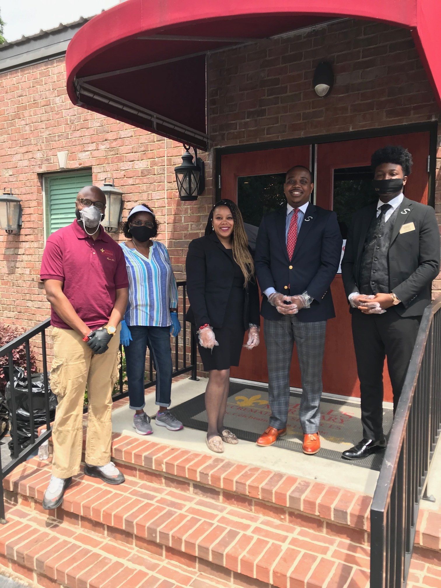Five people stand on a brick building's entrance steps, some wearing masks and gloves, posing for a photo.
