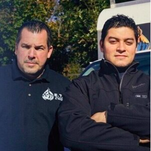 Two men are standing next to each other with their arms crossed and one has a flame logo on his shirt