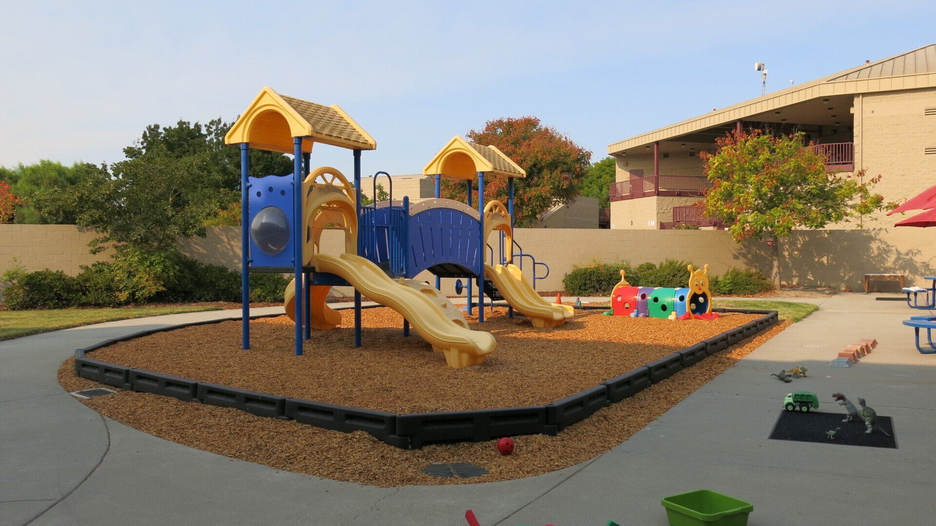A playground with a slide , swings , and a sandbox in front of a building.