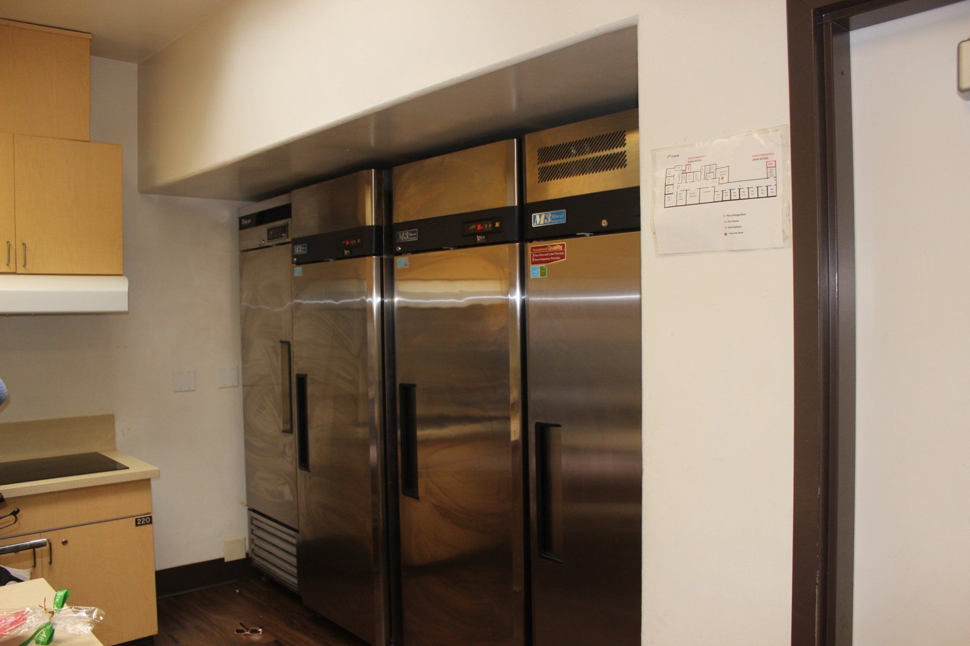 A kitchen with three stainless steel refrigerators and a sign on the wall.
