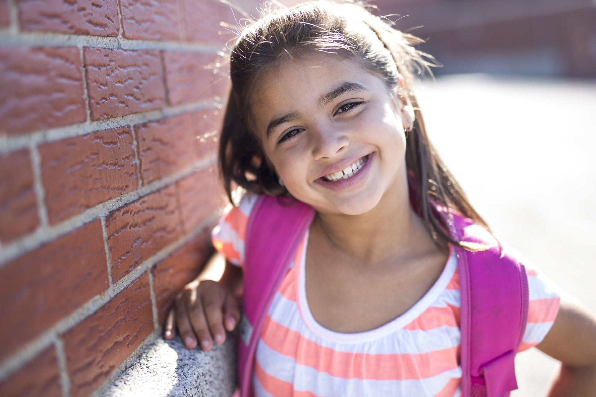 A little girl with a pink backpack is leaning against a brick wall and smiling.