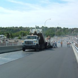 Asphalt paving on a bridge; dump truck unloading material with workers nearby under a blue sky.