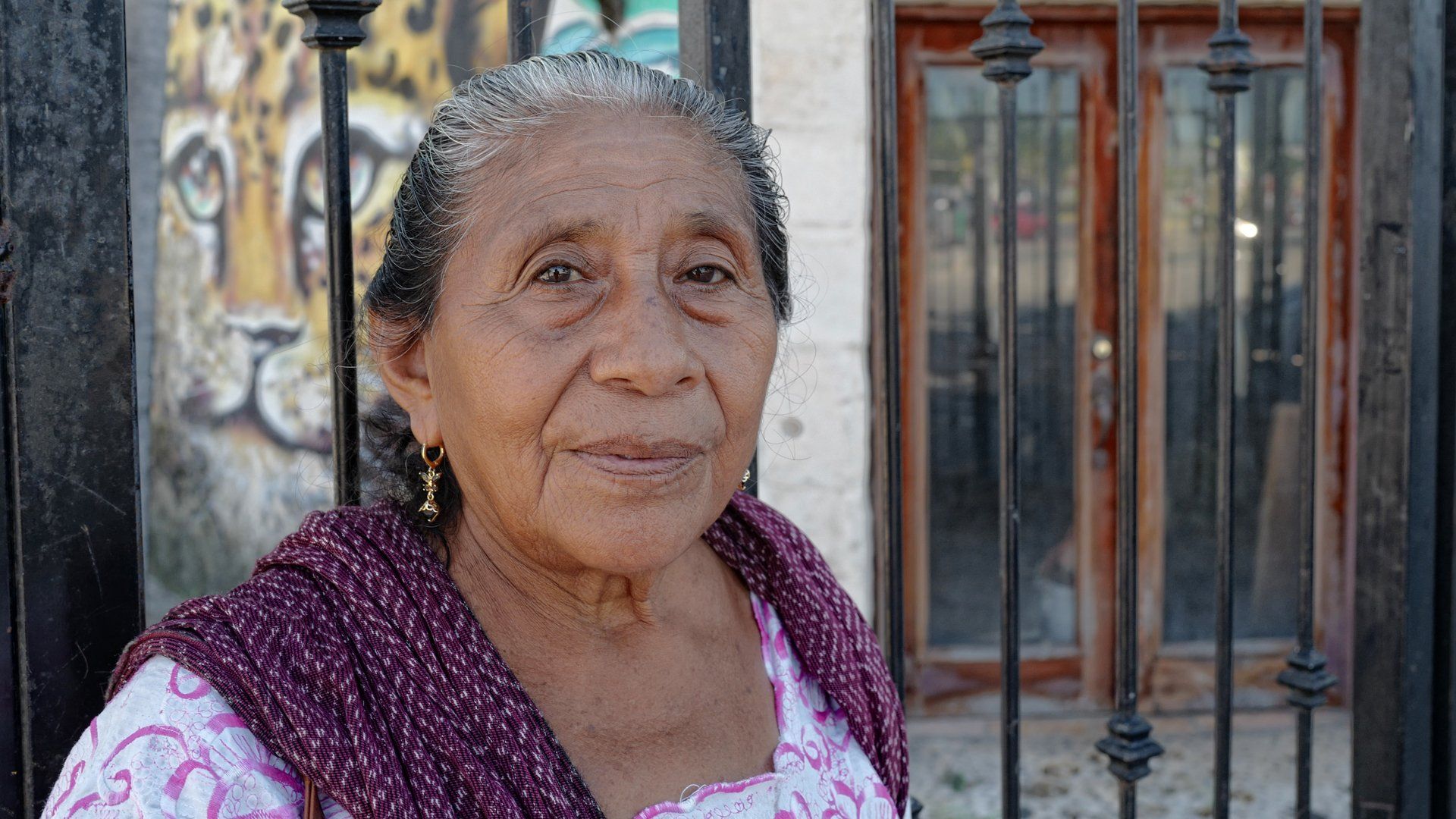 An elderly woman in a purple scarf is standing in front of a building.