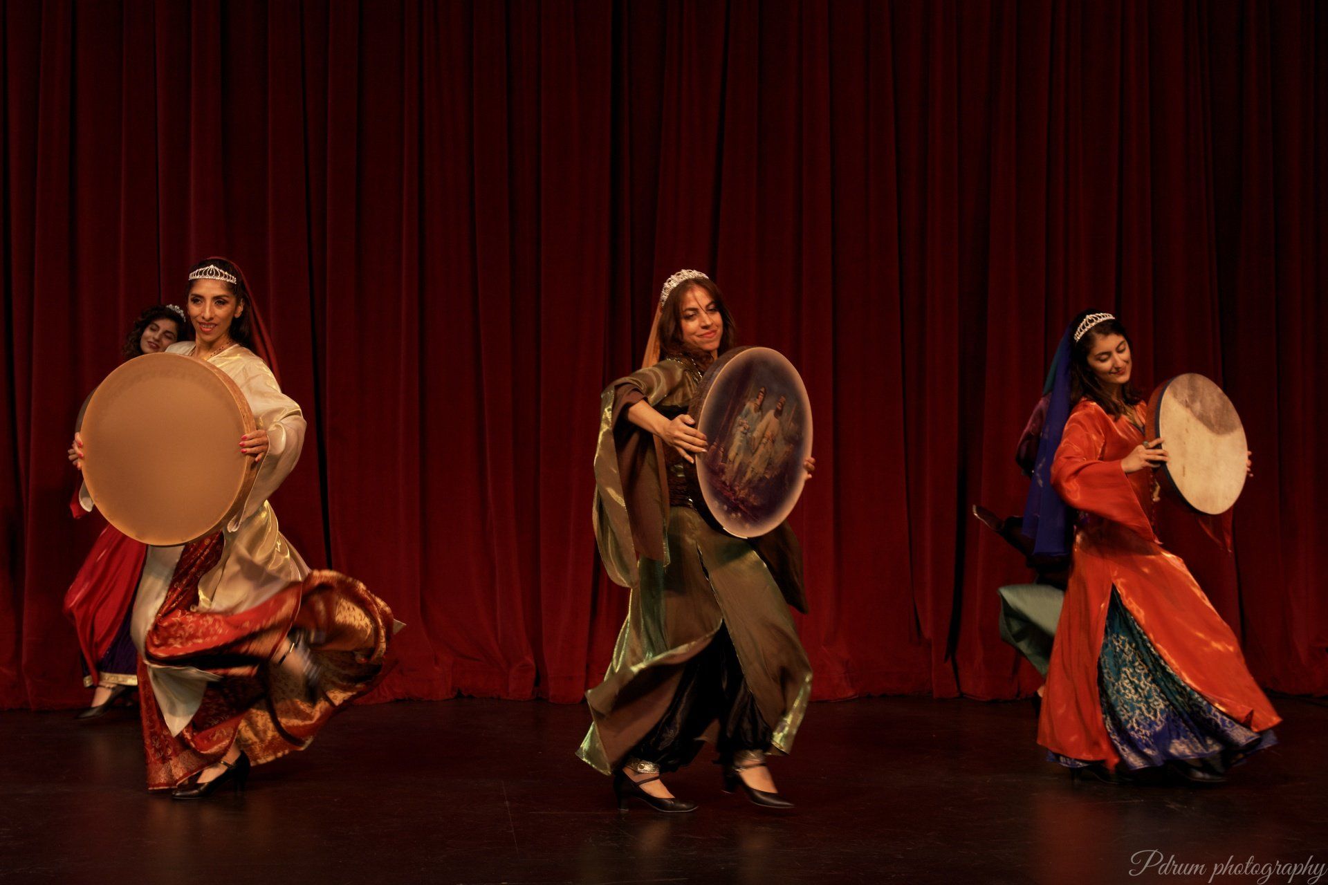 Three women are playing drums on a stage in front of a red curtain.