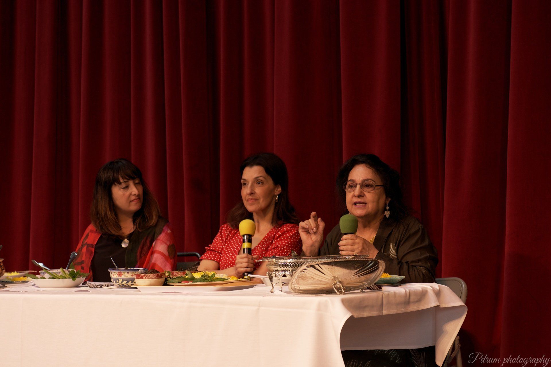 Three women are sitting at a table with microphones in front of a red curtain.