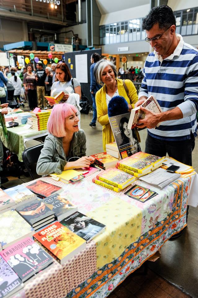 A woman with pink hair is sitting at a table with books on it.