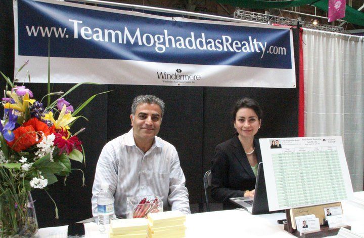 A man and a woman sit at a table in front of a team moghaddas realty banner