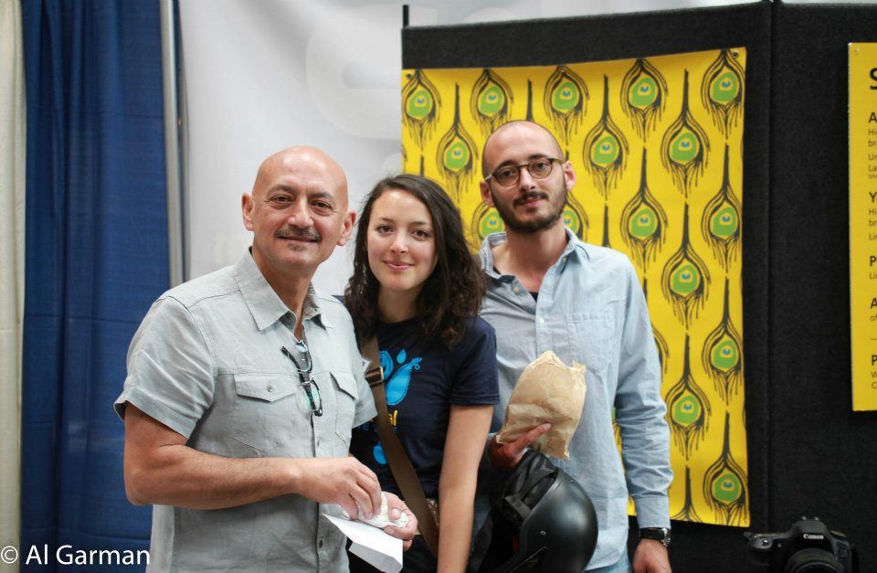 Three people are posing for a picture in front of a wall with peacock feathers on it.