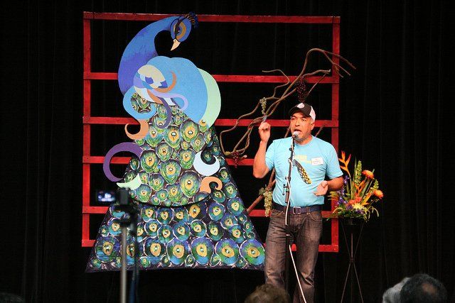 A man is standing in front of a microphone on a stage with a peacock in the background.