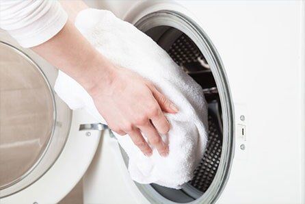 Woman Hands Loading Linen Into Washing Machine — Air-duct Cleaning in Deer Park, WA Person putting a white towel into a white washing machine.