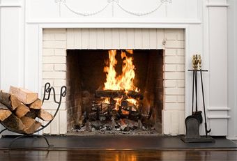 Chimney Cleaning — White Stone Frieplace in Deer Park, WA Fireplace with burning fire, brick surround, logs in rack on left, tools on right.