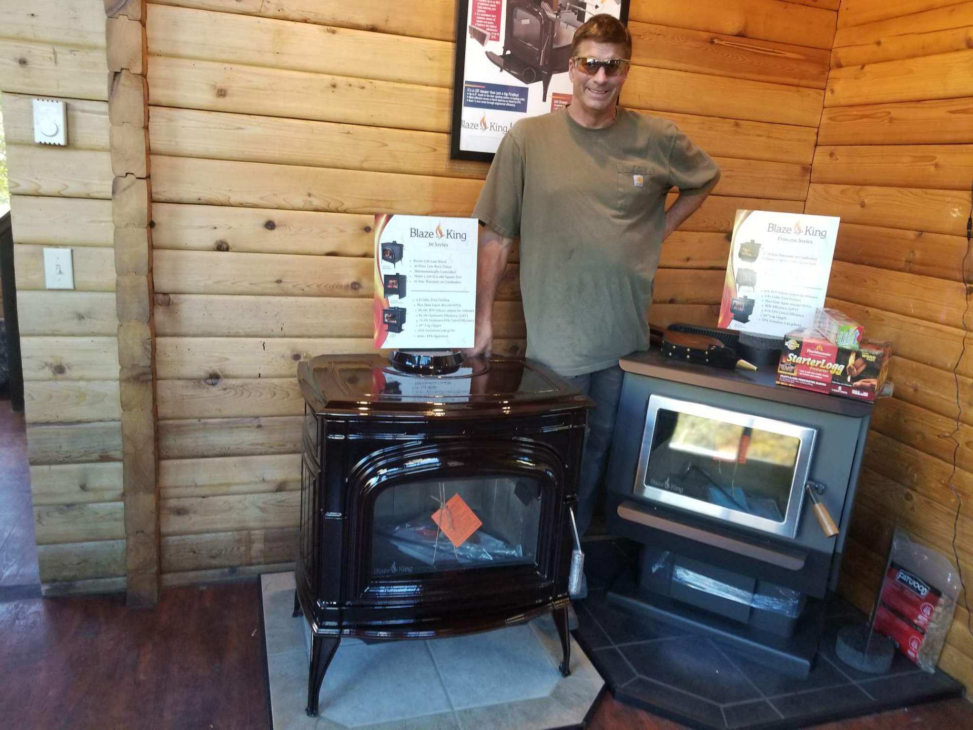 A man stands beside two wood-burning stoves in a wood-paneled room.