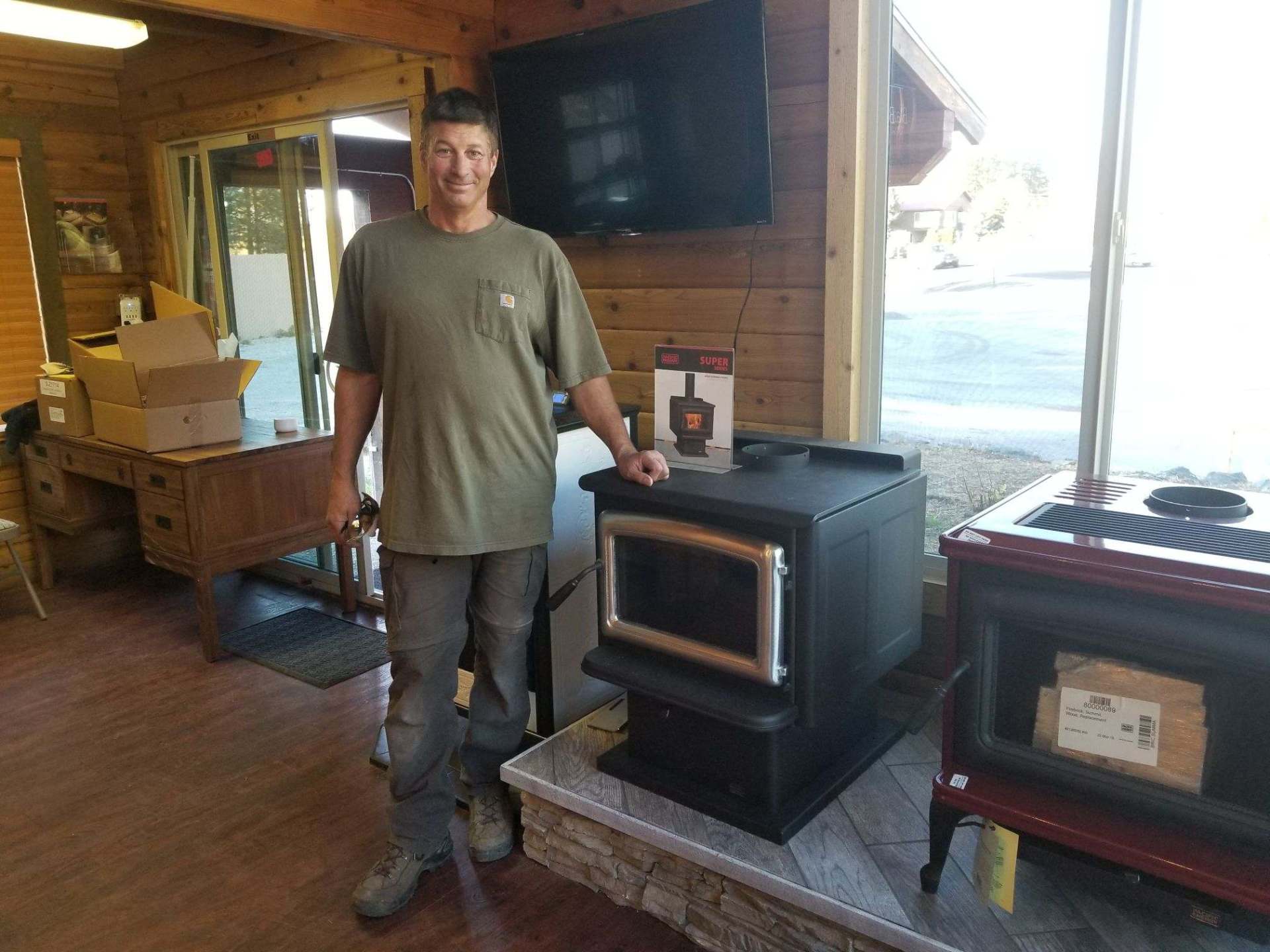 Man in a shop stands next to a black wood stove, with another red stove nearby.