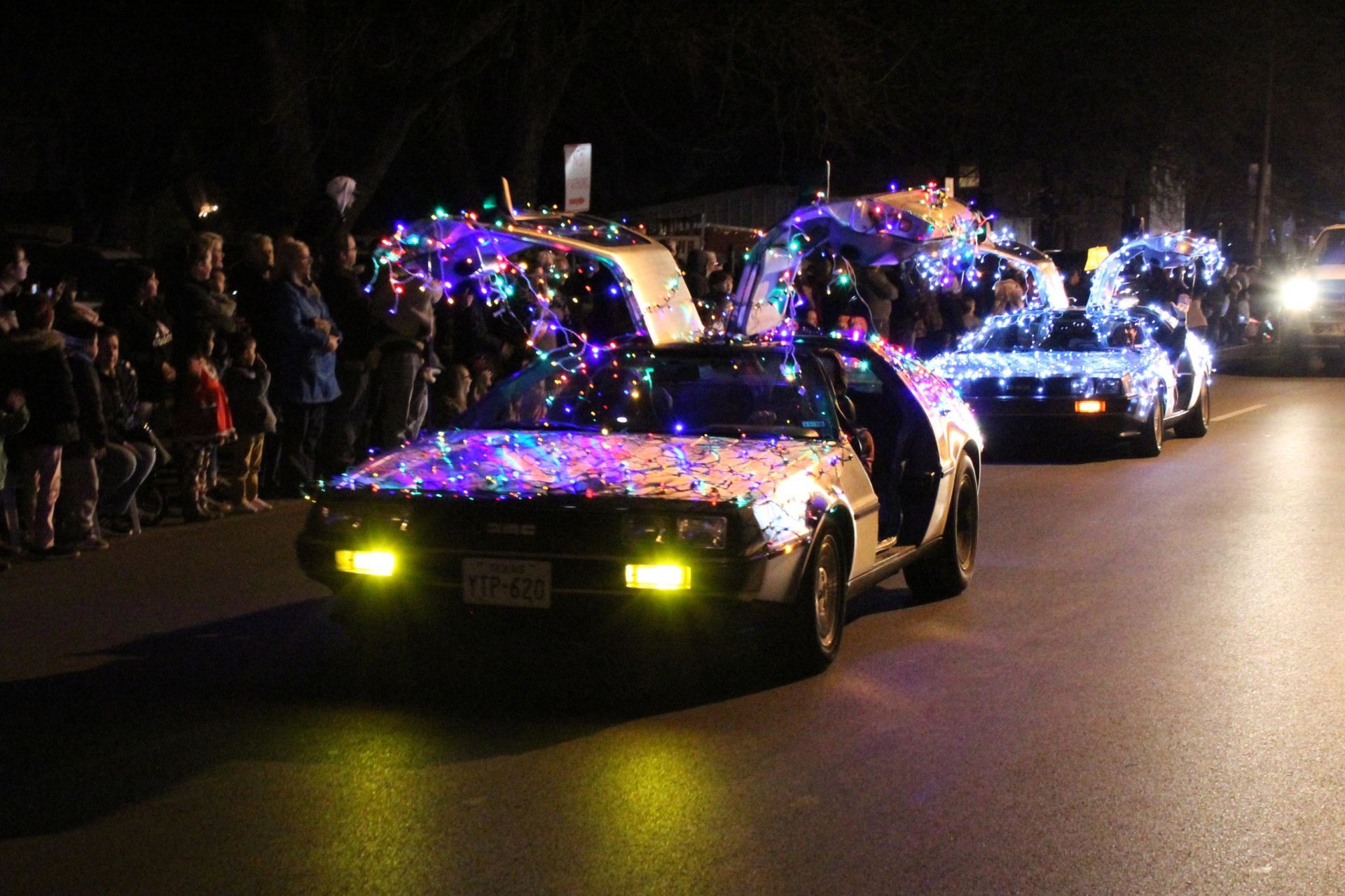 DeLorean cars decorated with colorful lights in a night parade, doors open, with spectators on the sidelines.