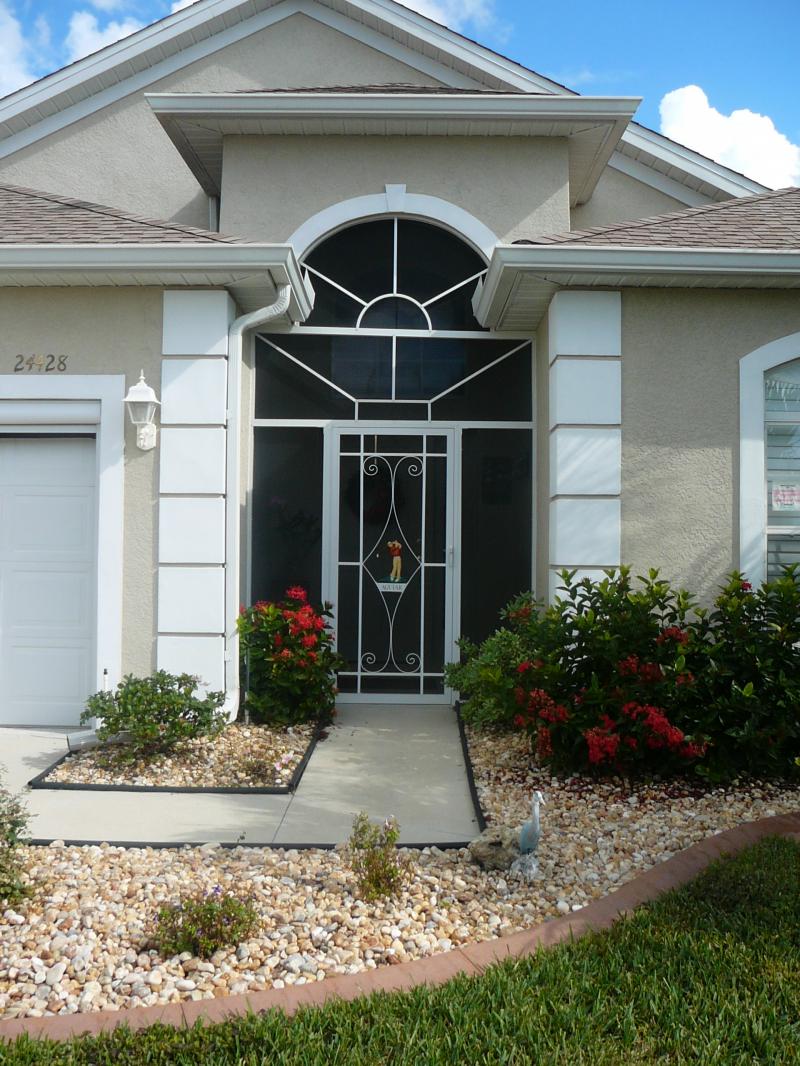 Tan house with white trim, arched screen door, small front yard with bushes.