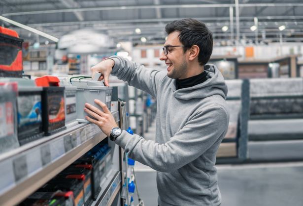 Una persona sorridente con indosso una felpa grigia con cappuccio osserva le batterie per auto su uno scaffale di un negozio.