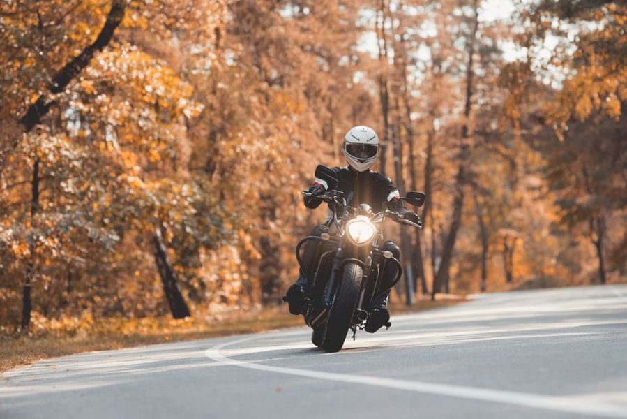 Un motociclista con il casco percorre in sella a una moto scura una strada tortuosa fiancheggiata da alberi autunnali.