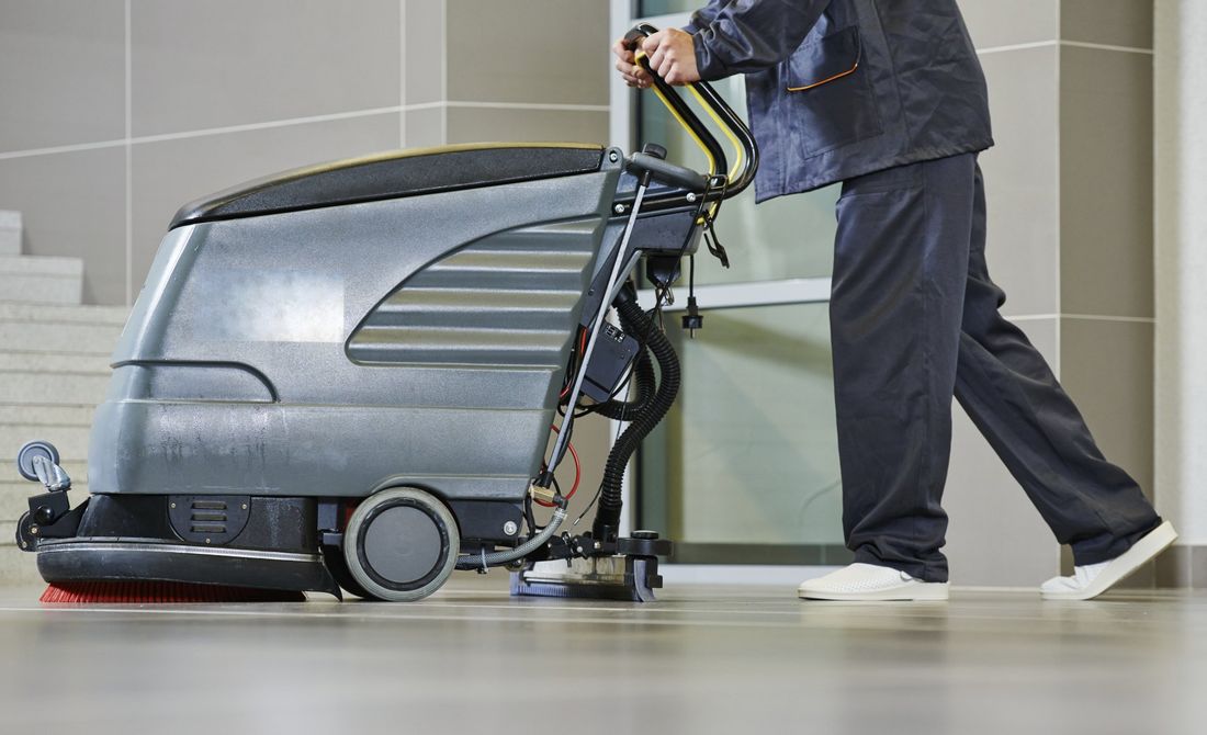 Una persona in uniforme scura utilizza una lavasciuga pavimenti a spinta per pulire un corridoio interno piastrellato.