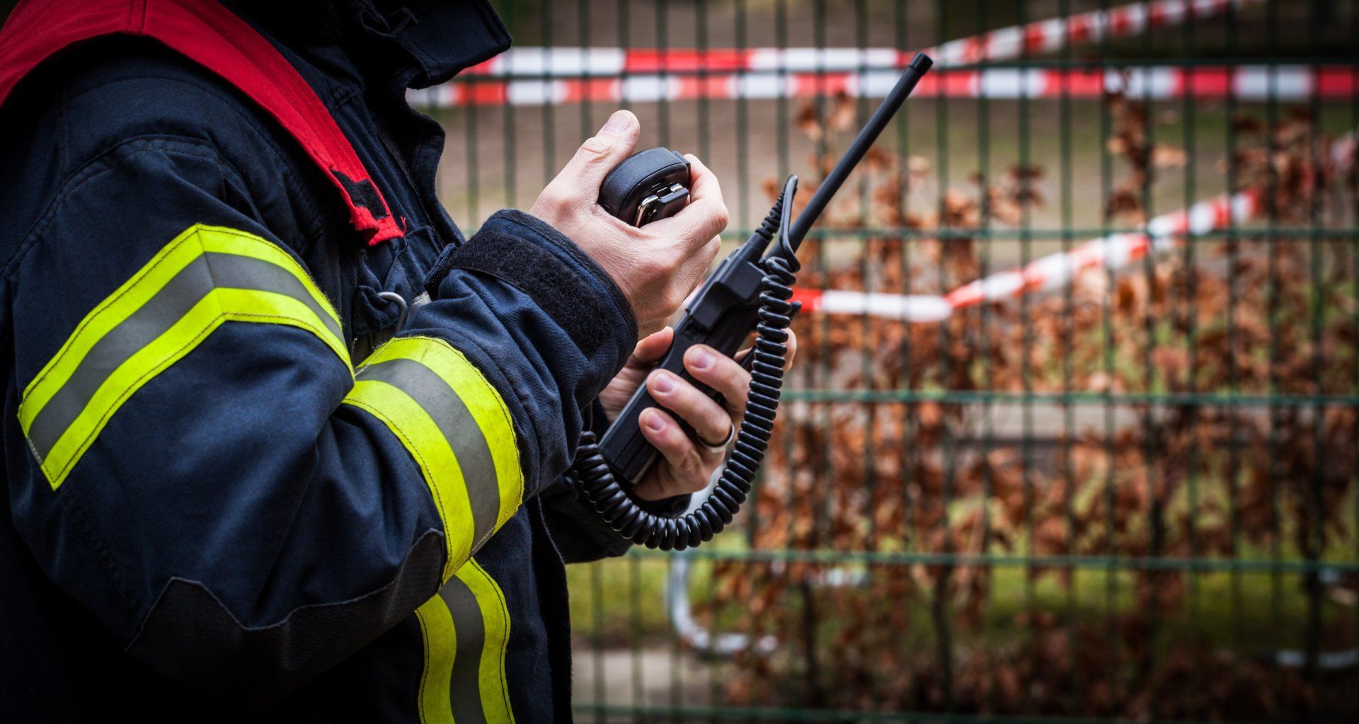 A fireman is talking on a walkie talkie in front of a fence.