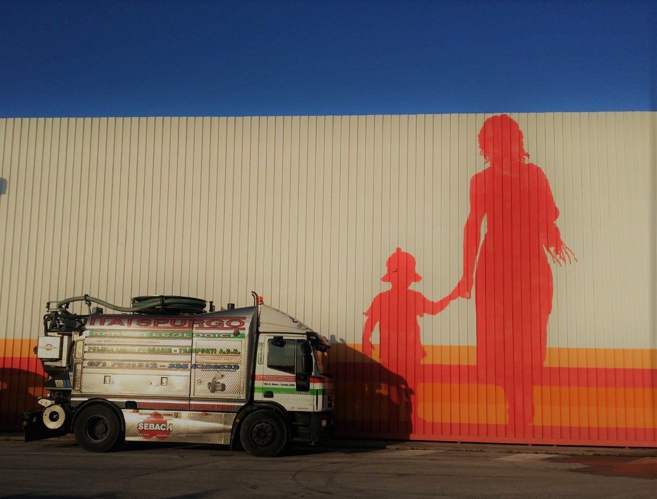 Un camion è affiancato da un murale raffigurante un adulto e un bambino in rosso che si tengono per mano sotto un cielo blu.