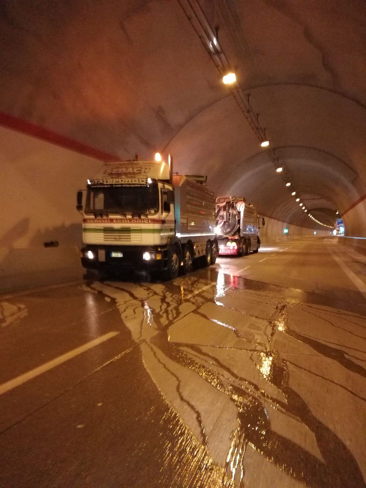 Due camion in una galleria bagnata, con i riflessi della strada e le luci del soffitto che illuminano la scena.