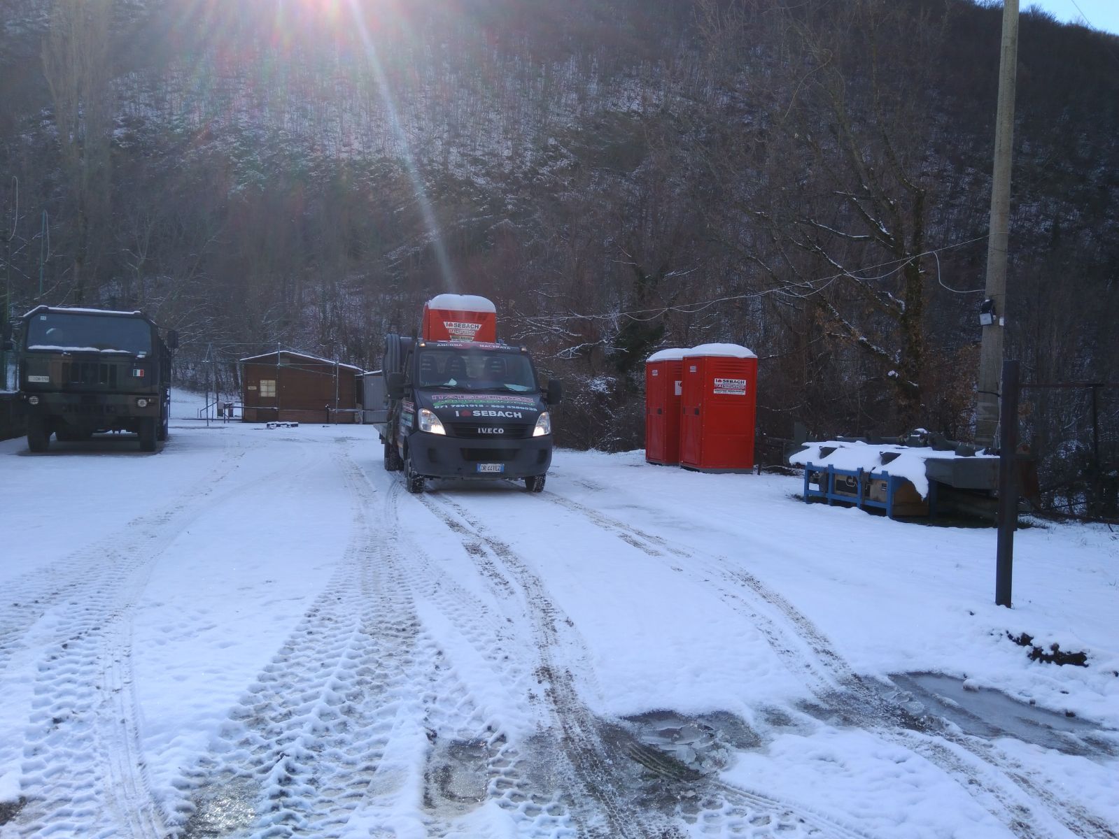 Strada innevata con veicoli, tra cui un furgone scuro e bagni chimici rossi, sotto un cielo luminoso