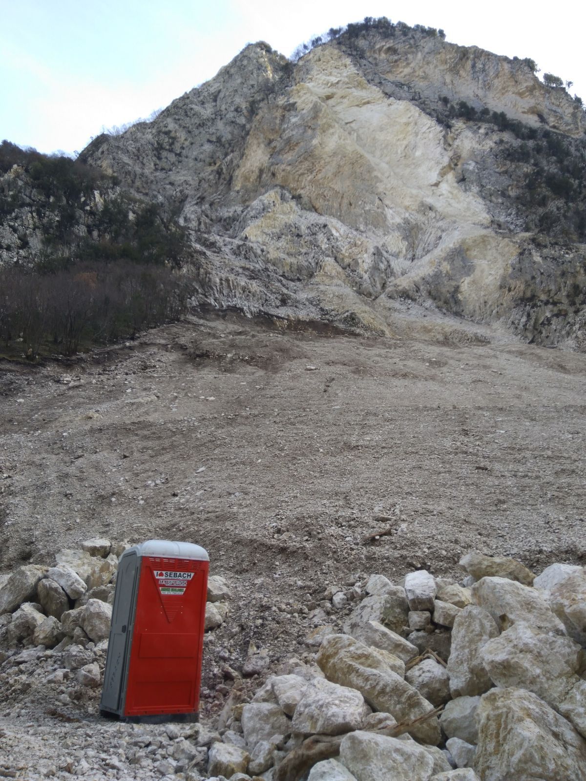 Toilette portatile rossa ai piedi di una frana rocciosa su un pendio montuoso brullo.