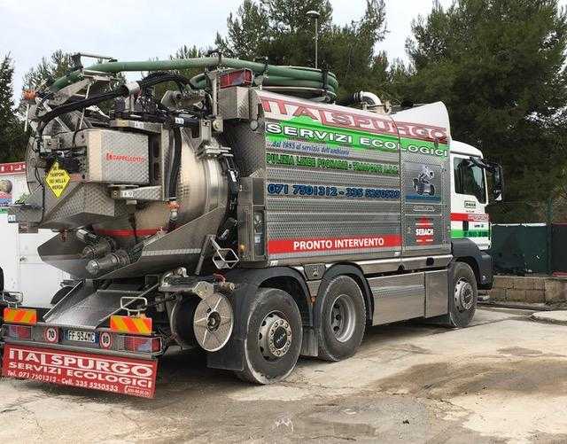 Un grosso camion aspiratore grigio con tubi e serbatoi parcheggiato all'aperto su un terreno sterrato.