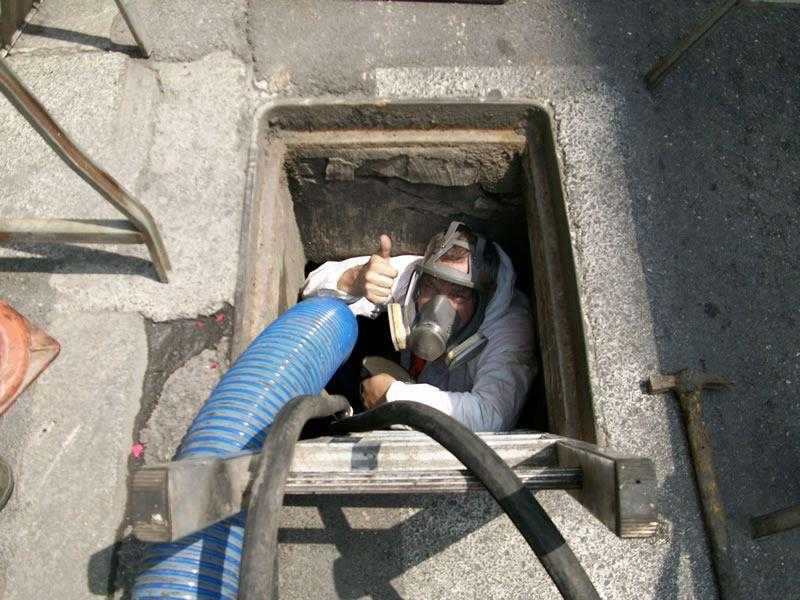 Un operaio all'interno di un tombino con tubi e attrezzi, visto dall'alto.
