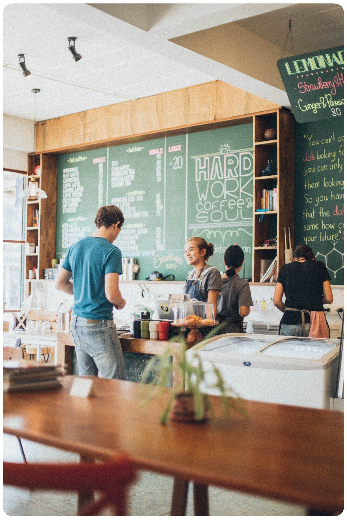 Inside a cafe, a customer orders from a counter where two employees stand in front of a chalkboard menu.