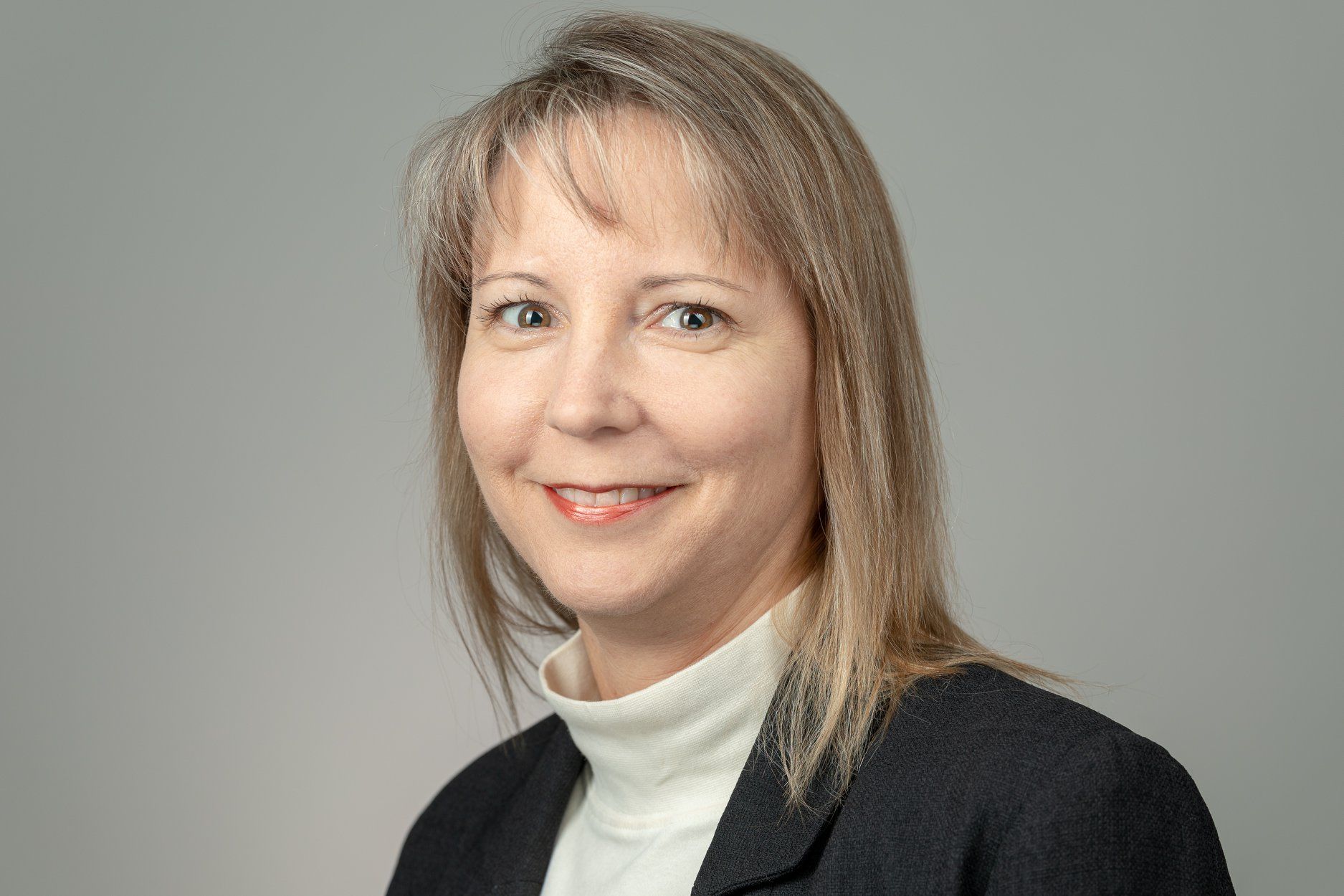 Woman with light hair smiles, wearing a black blazer and white turtleneck. Gray background.