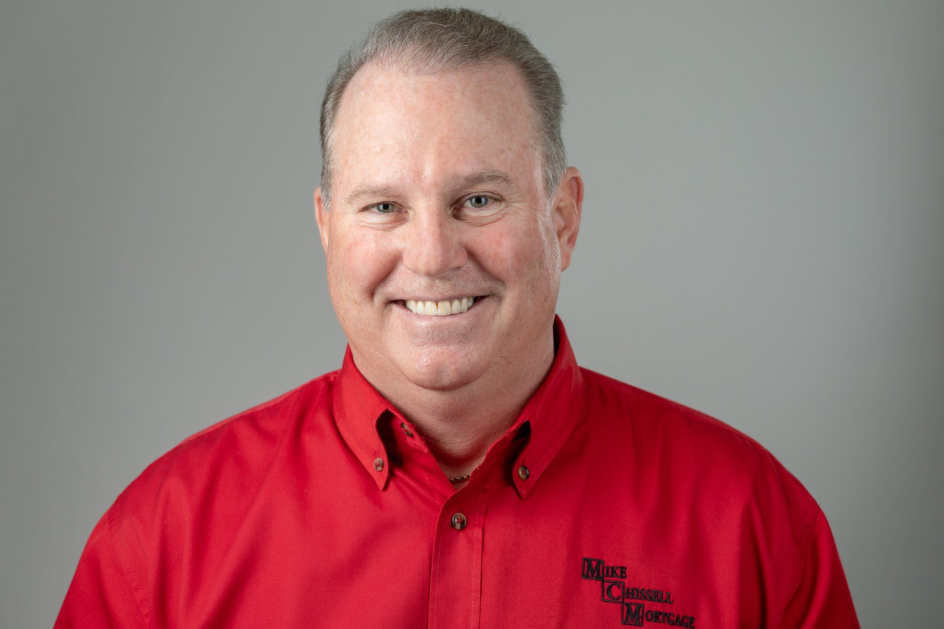 Man in red shirt smiles at the camera, with a logo on shirt, against a gray background.