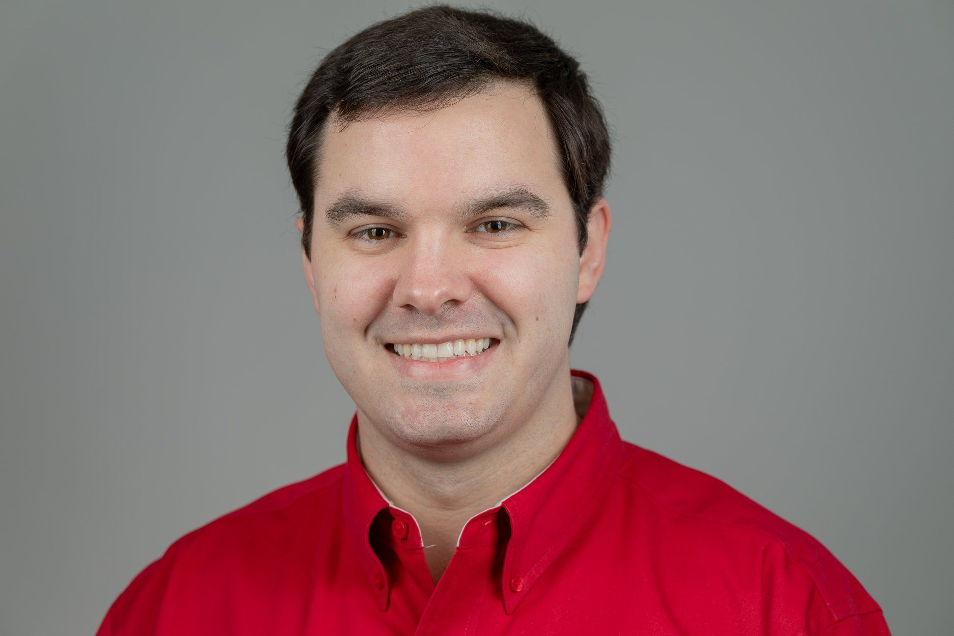 Man wearing a red shirt smiles at the camera against a gray background.