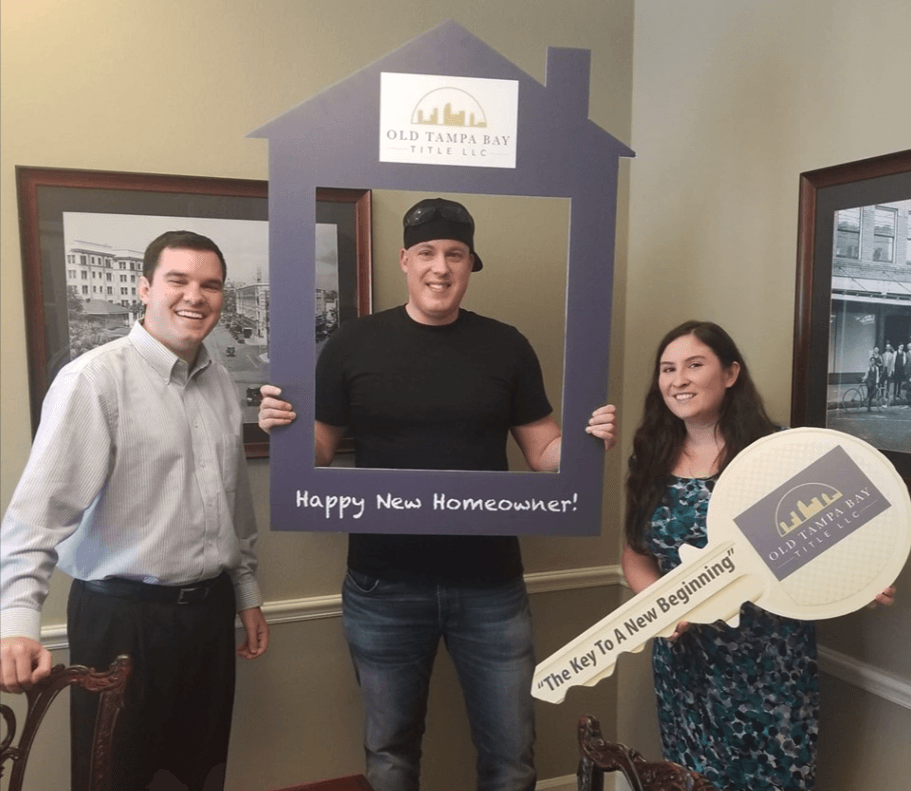 Three people pose for a photo. A man in the center holds a house-shaped frame.